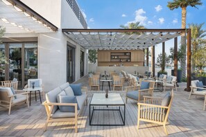 Patio esterno con disposizione di posti a sedere, un tavolo centrale e un'area cucina visibile attraverso le vetrate.