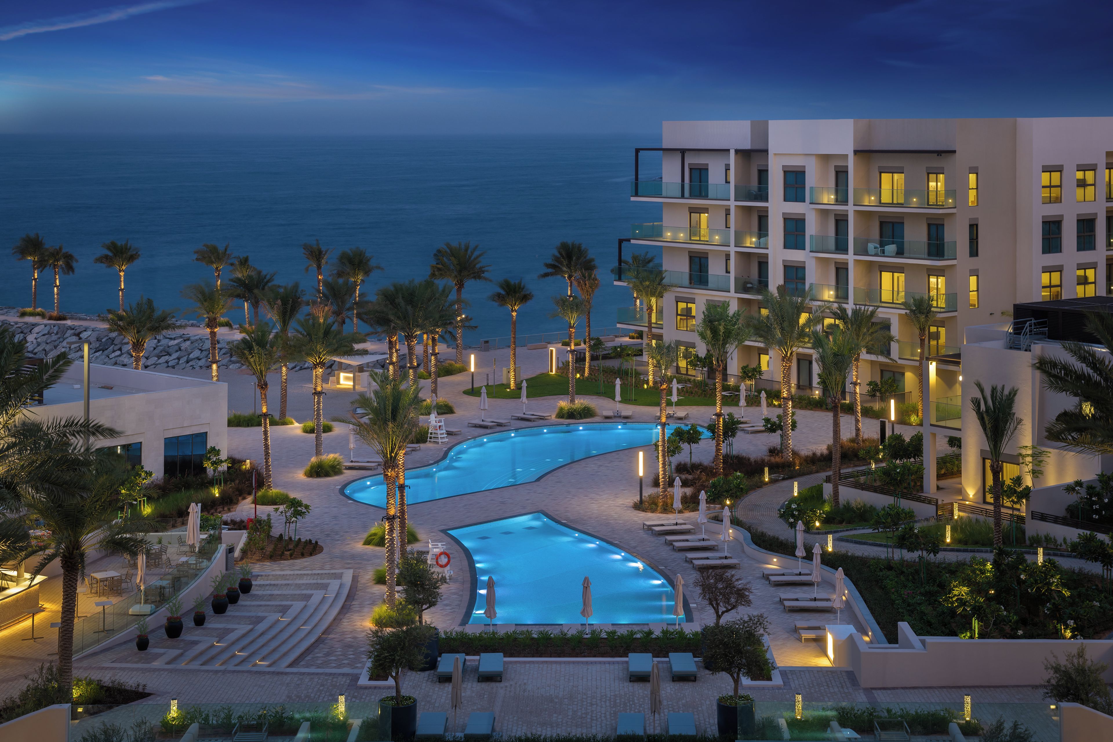 A resort pool area with a curved pool, palm trees, and a view of the ocean.