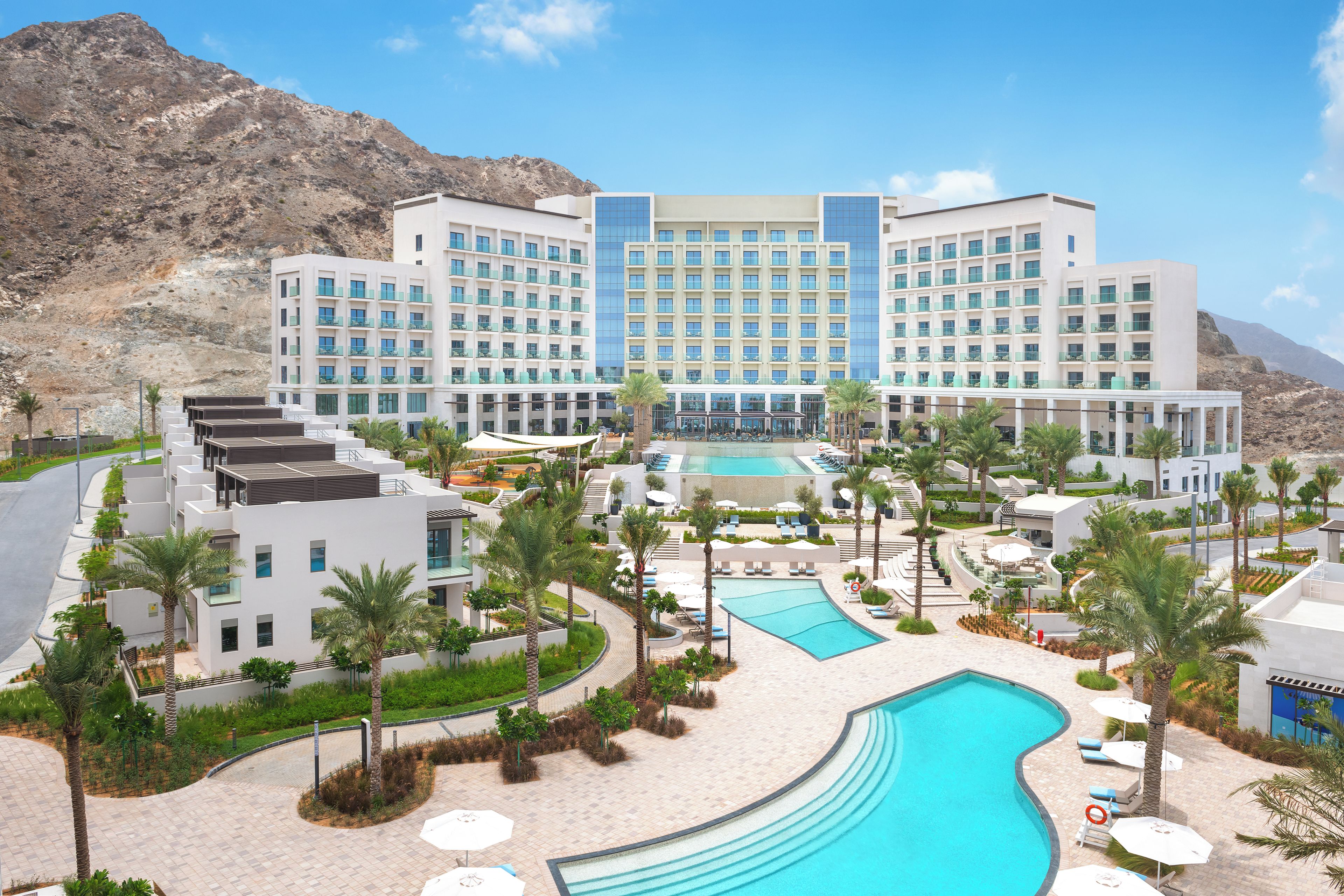 A hotel with a pool, palm trees, and a mountainous backdrop.