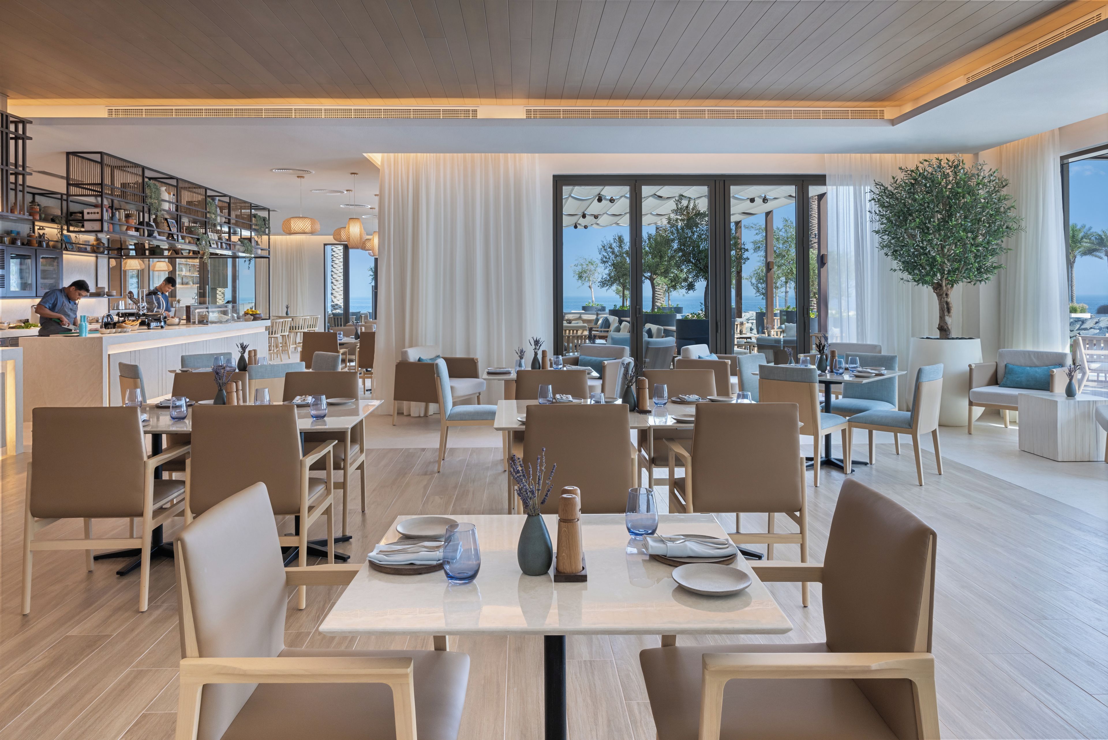 Un amplio comedor con varias mesas y sillas, una zona de cocina con personal, y grandes ventanales que ofrecen vistas al mar.