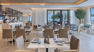 Un amplio comedor con varias mesas y sillas, una zona de cocina con personal, y grandes ventanales que ofrecen vistas al mar.