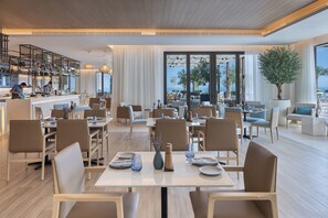 Een ruime eethoek met meerdere tafels en stoelen, een keukenruimte met personeel, en grote ramen met uitzicht op zee.