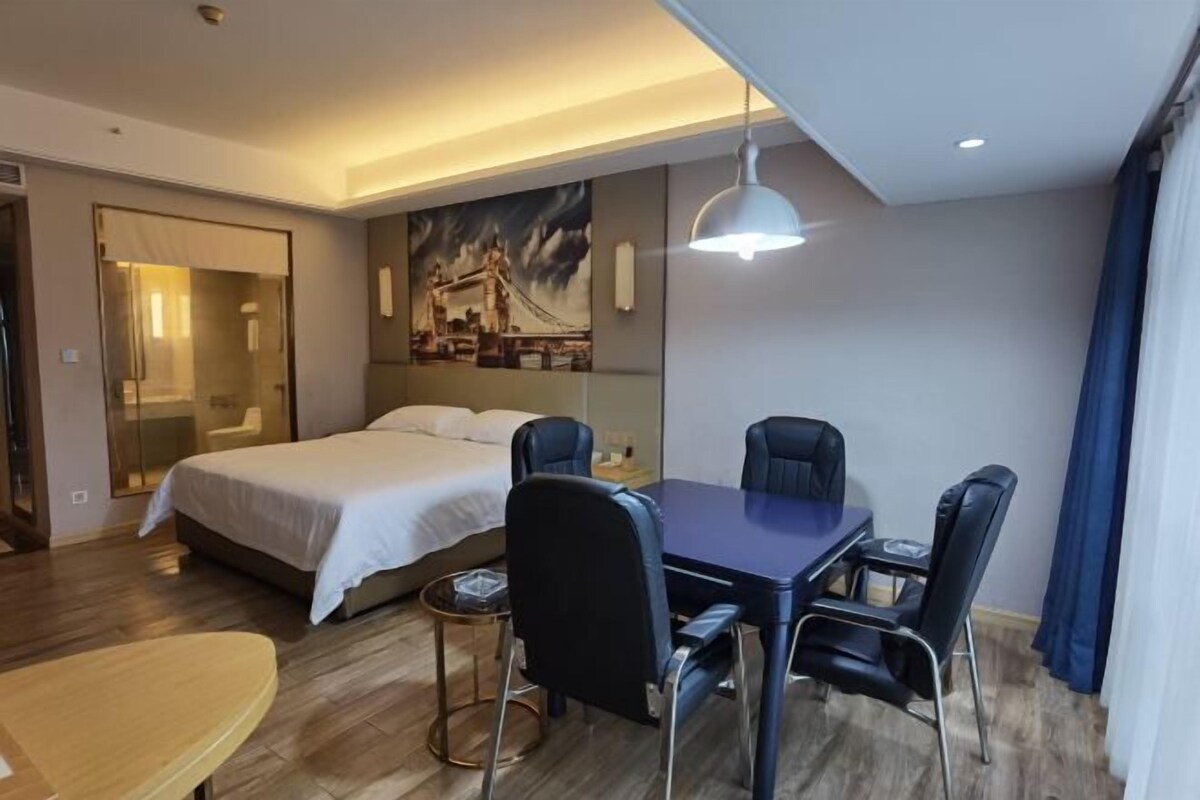 A hotel room with a bed, a dining table with chairs, a painting on the wall, and a ceiling light.