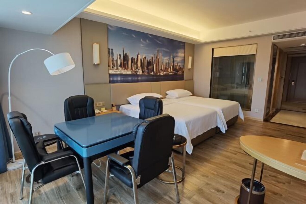 A hotel room with a bed, a desk with chairs, a lamp, and a painting of a cityscape on the wall.