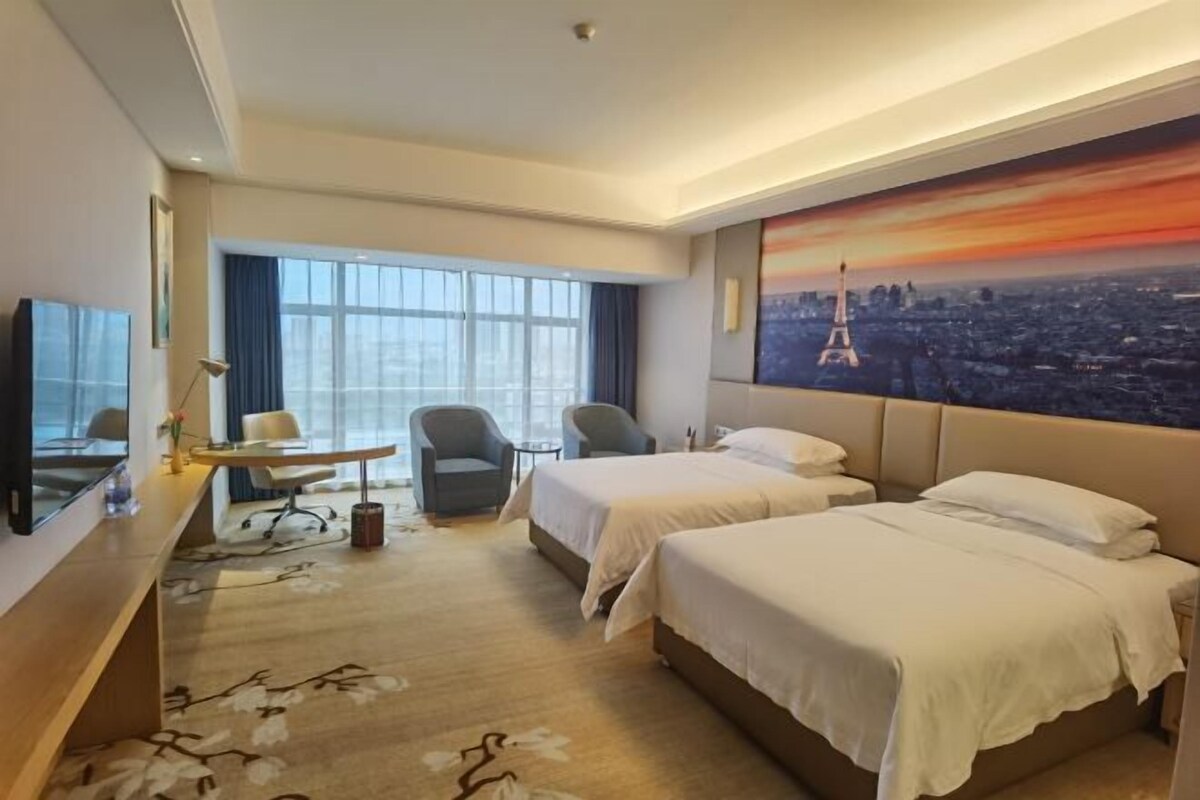 A hotel room with two beds, a desk, a chair, a TV, and a large window with a cityscape view.