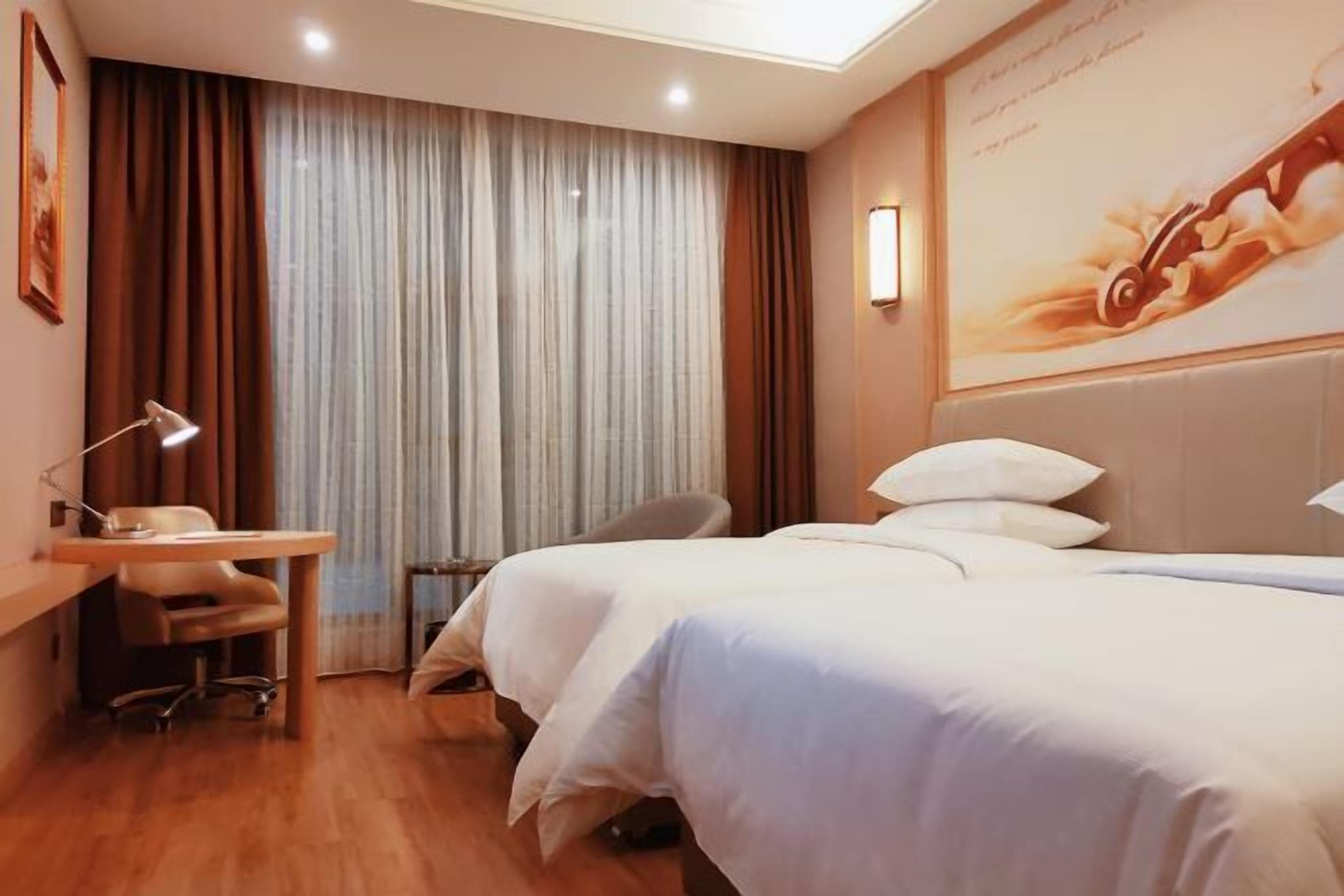 A hotel room with two beds, a desk with a chair, a lamp, and a large poster on the wall.