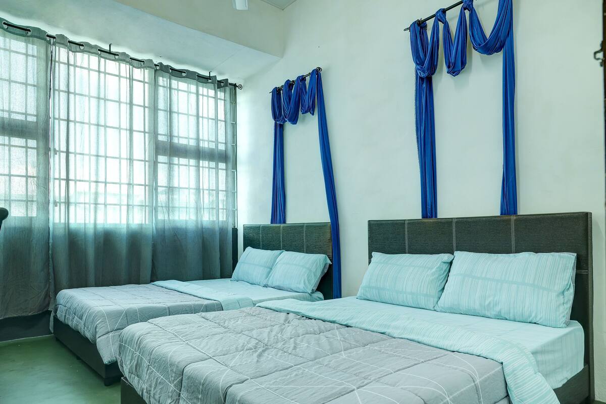 A hotel room with two beds, a large window with sheer curtains, and blue drapes on the window frames.