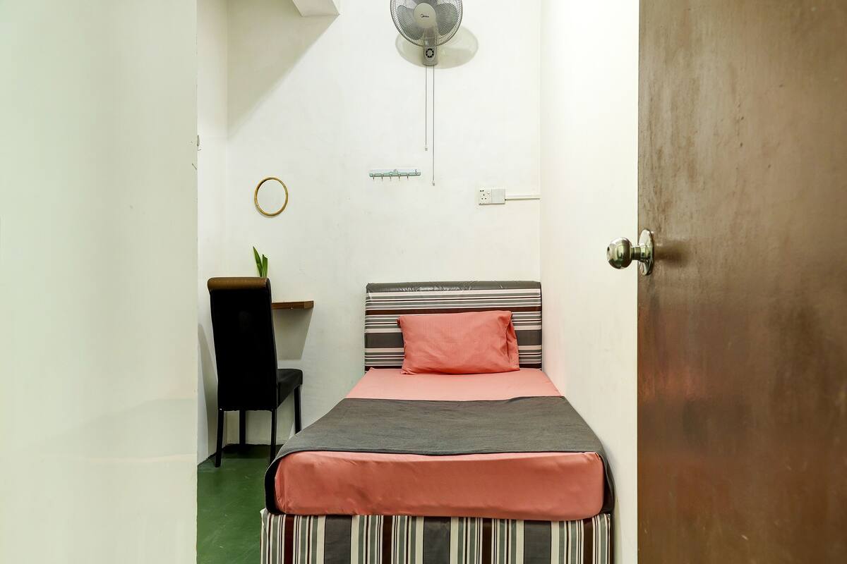 A small, neatly arranged bedroom with a bed, a chair, a desk, and a wall-mounted fan.