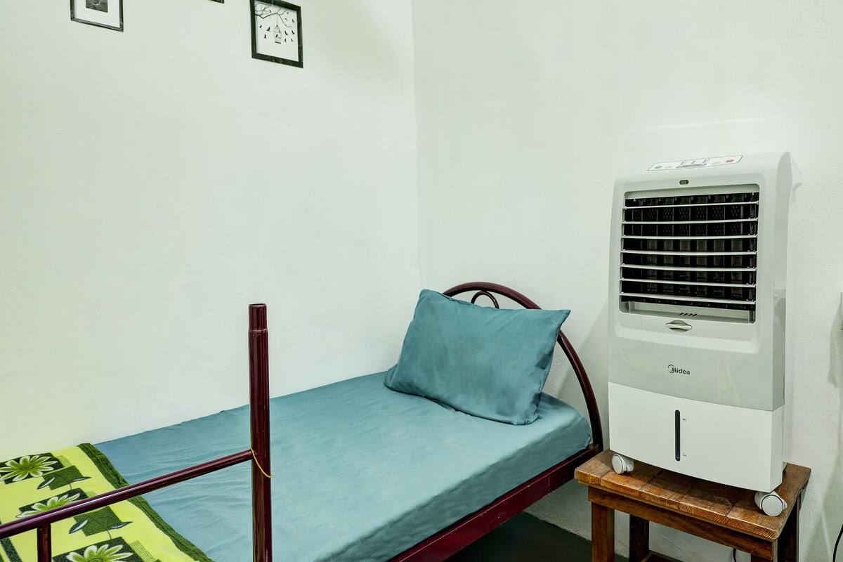 A single bed with a blue mattress and a pillow, a wooden frame, a small bedside table with a white device, and a wall-mounted air conditioner.