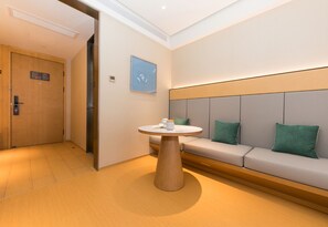 Hotellihuone, jossa on pyöreä pöytä, tyynyillä varustettu sohva, ovi ja seinälle ripustettu taulu.