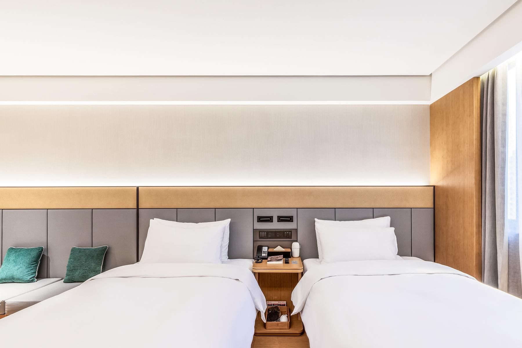 A hotel room with two beds, a bedside table, a lamp, and a window with curtains.