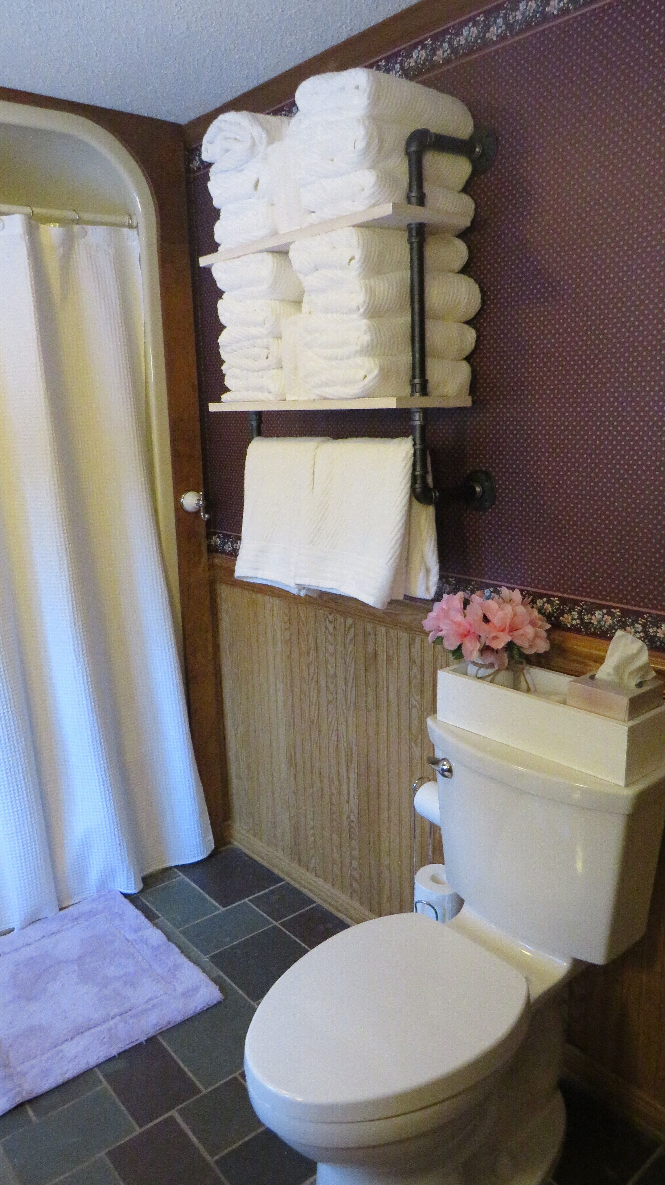 A bathroom with a toilet, a towel rack with neatly folded towels, a toilet paper holder, and a small vase with flowers on the sink.