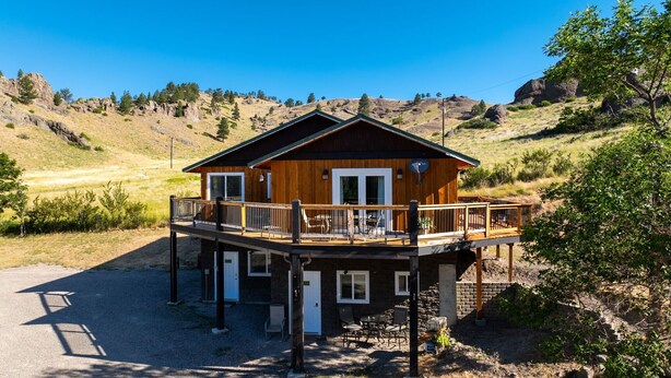 A two-story wooden cabin with a balcony, surrounded by a gravel driveway and a mountainous landscape.