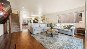 Ein geräumiges Wohnzimmer mit einem hellgrauen Ecksofa, einem Holztisch und einem gemusterten Teppich. Eine Treppe mit Holzbodenstufen führt in den Hintergrund zu einer Küchenzeile.