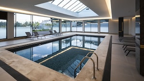 A modern indoor swimming pool with clear glass walls, lounge chairs, and a skylight.