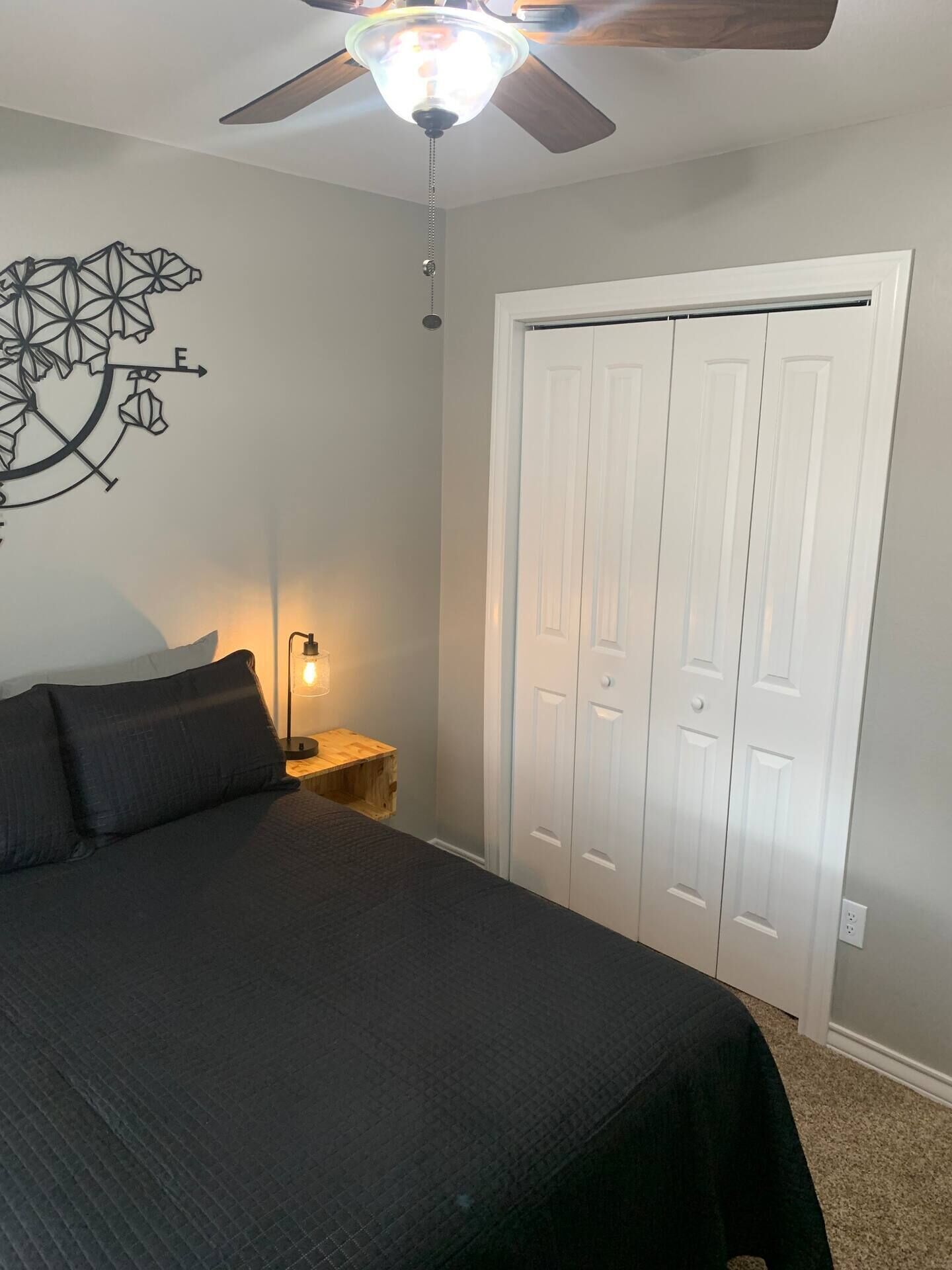 A bedroom with a bed, a ceiling fan, a wall art piece, a nightstand with a lamp, and a closet.