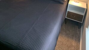 Uma cama bem arrumada com um edredom escuro, duas mesas de cabeceira com luminárias e uma parede lisa.