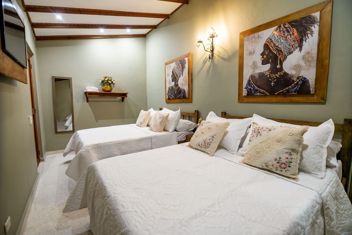 A hotel room with two beds, a small shelf with towels, a wall-mounted light, and two framed artworks.