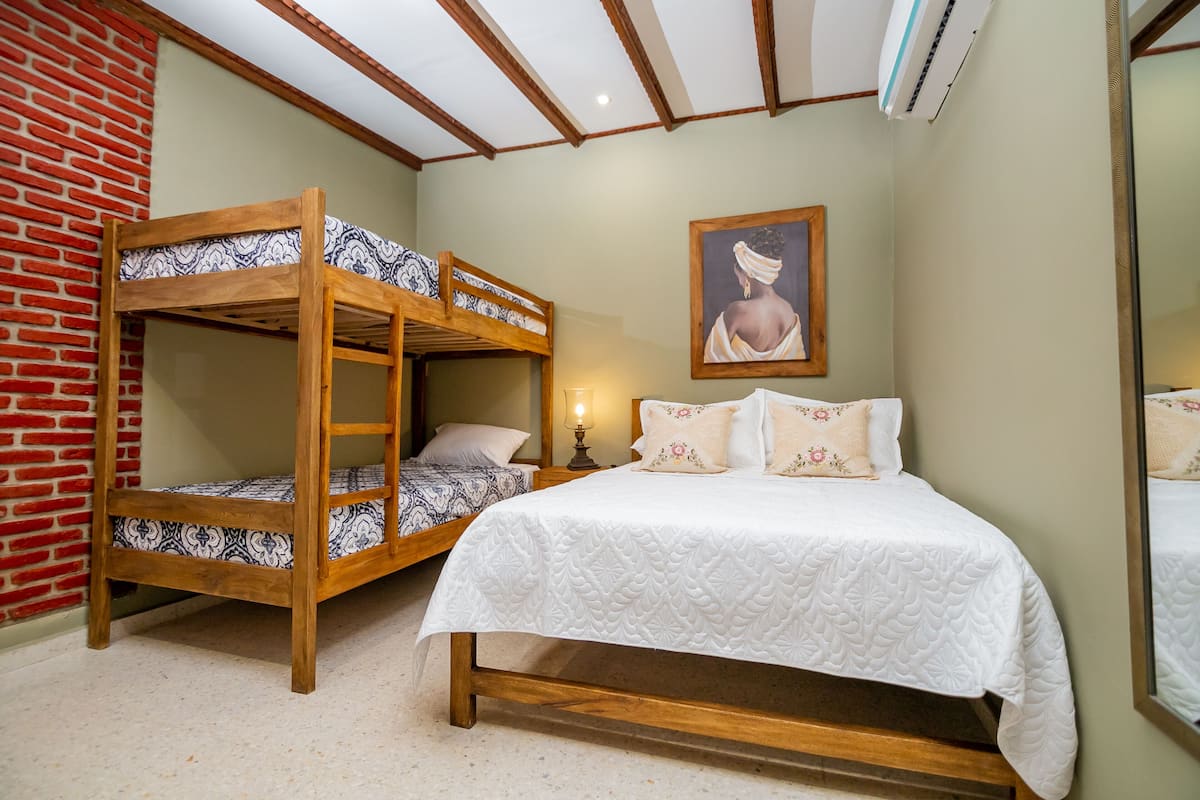 A room with a bunk bed, a single bed, a framed picture on the wall, and a nightstand with a lamp.