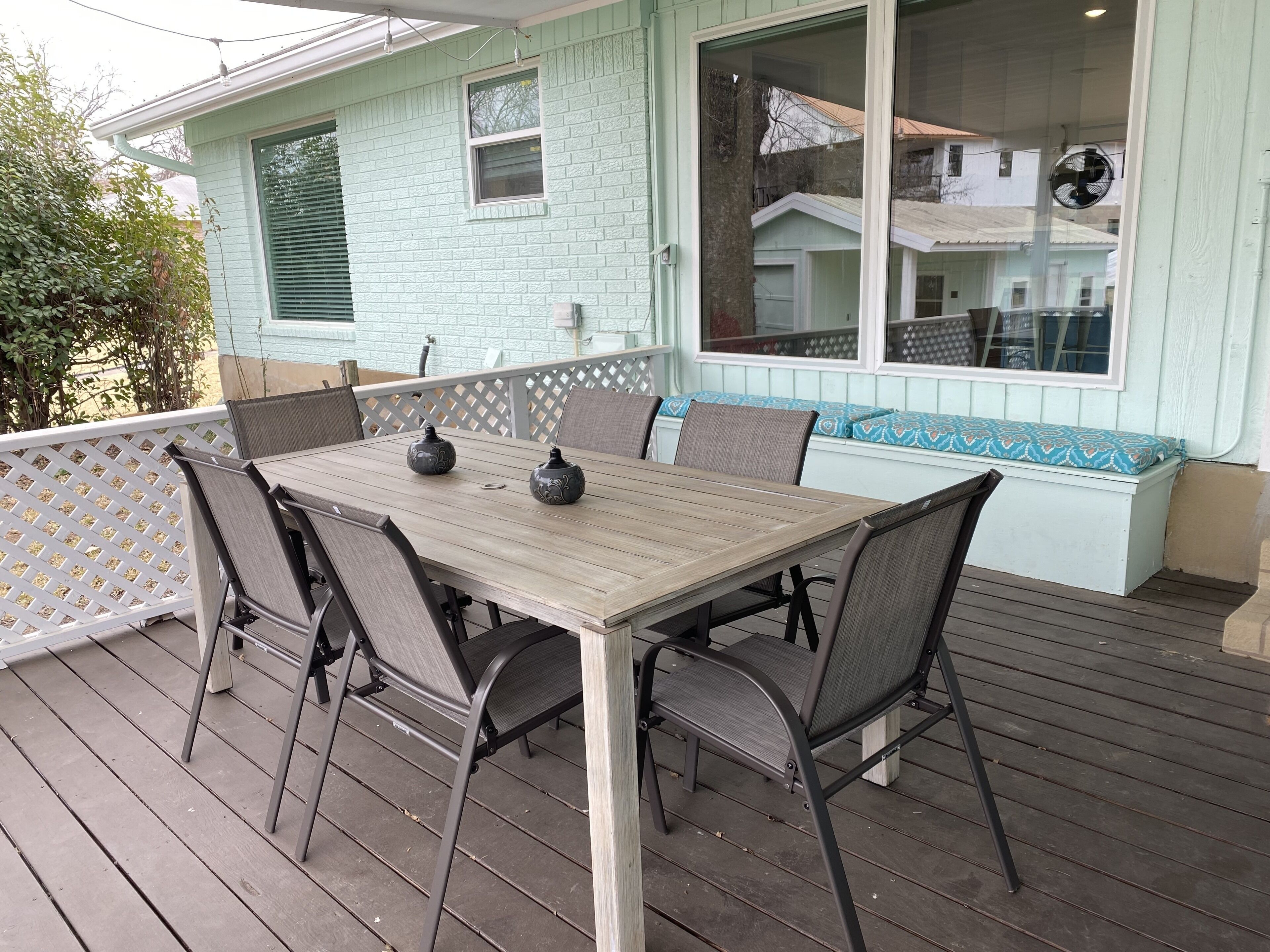 A wooden deck with a table and chairs, a white lattice fence, and a light blue building with windows and a fan.