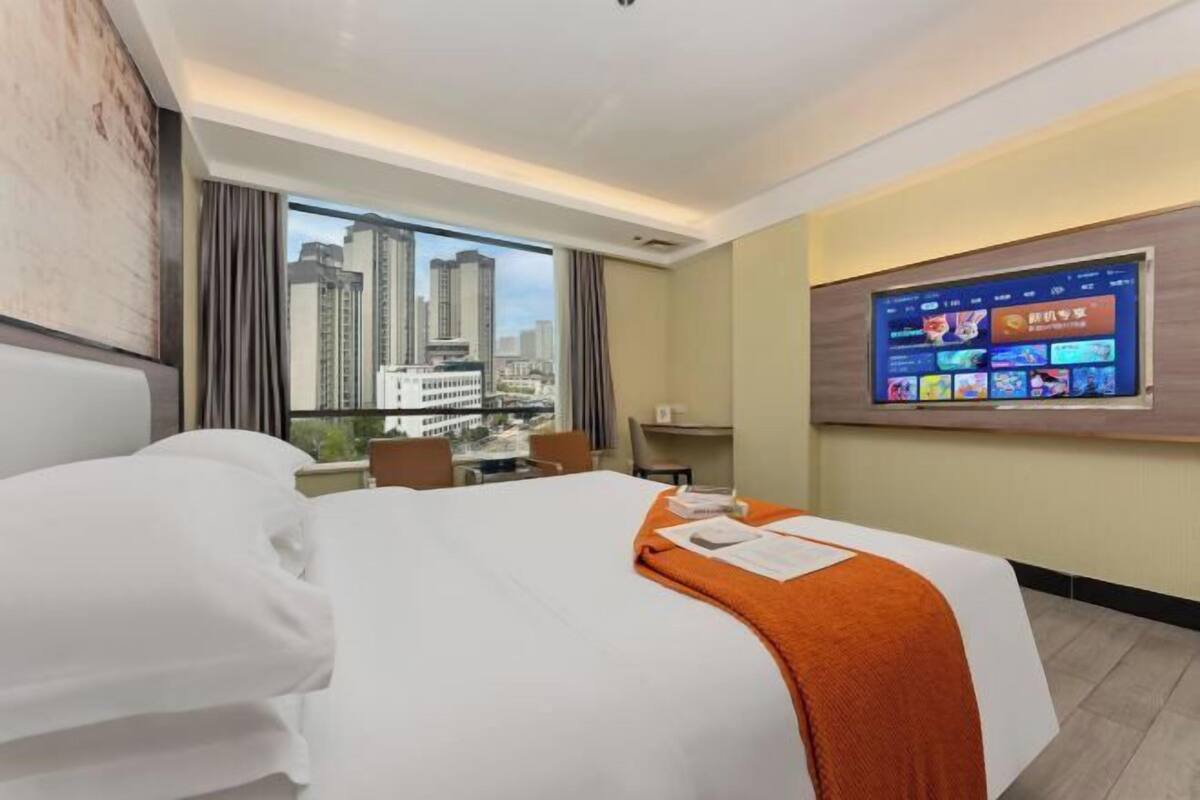 Habitación de hotel con cama, televisor montado en la pared, ventana con vista a la ciudad y escritorio con silla.