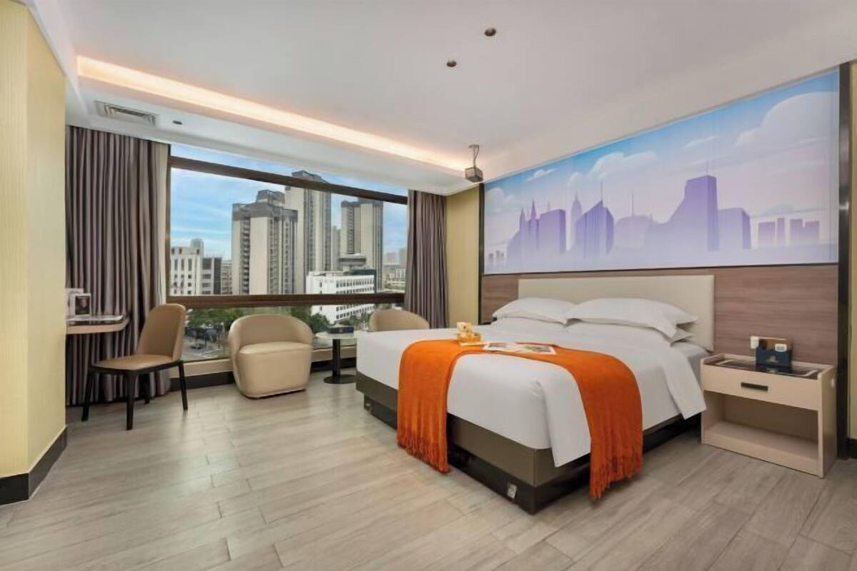 Habitación de hotel con cama, escritorio, dos sillas, mesita de noche y una ventana grande con vistas a la ciudad.