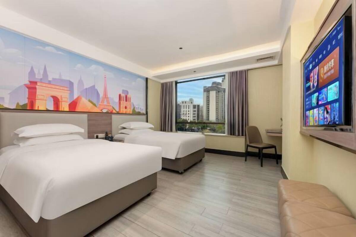 Habitación de hotel con dos camas, un ventanal grande con vistas a la ciudad, un televisor montado en la pared y un mural con monumentos famosos.
