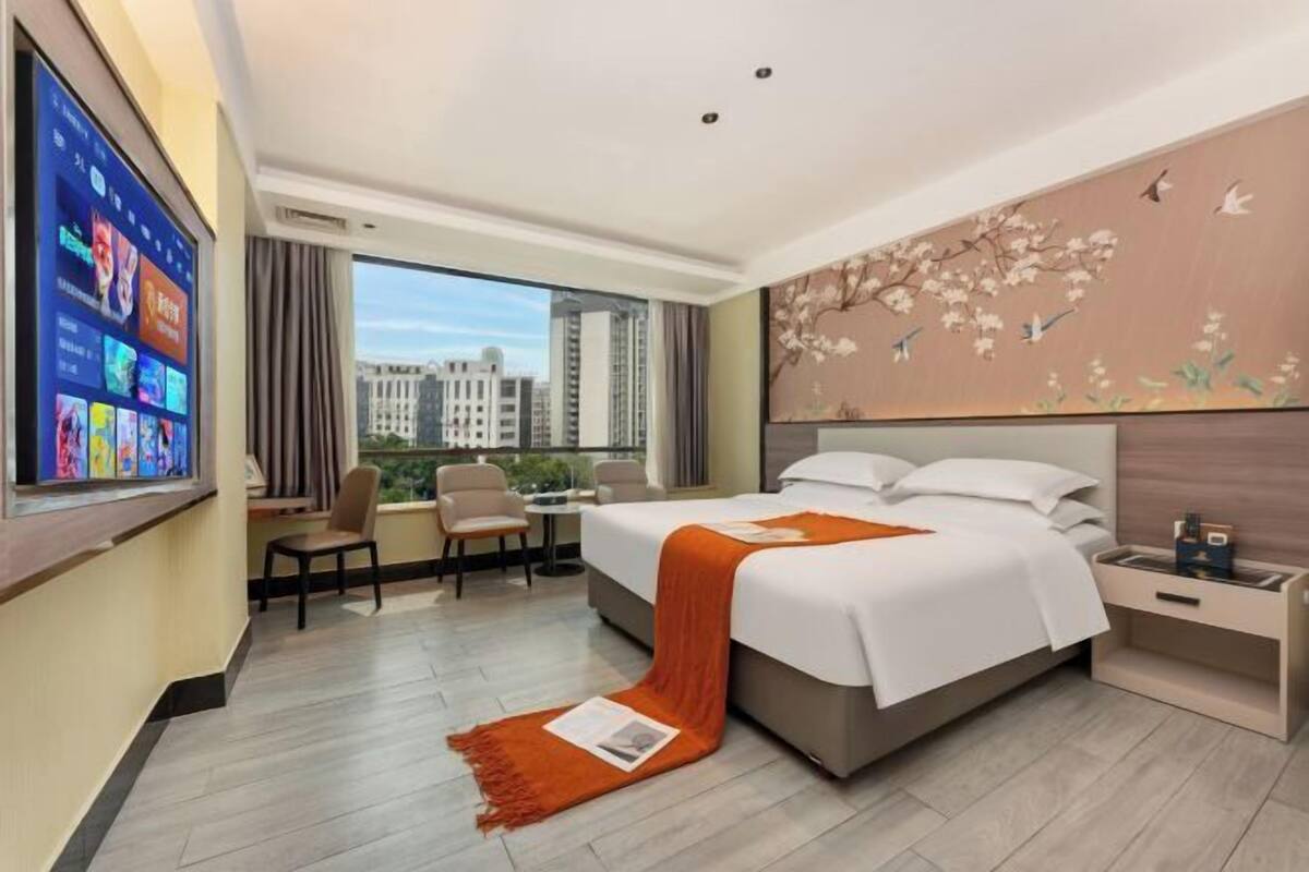 Habitación de hotel con cama, escritorio, silla, televisión y una ventana grande con vistas a la ciudad.