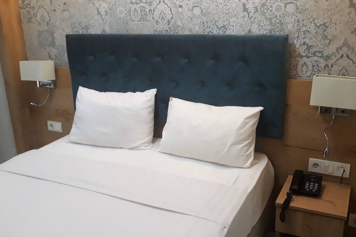 A hotel room with a bed, two white pillows, a blue headboard, two bedside lamps, and a bedside phone.