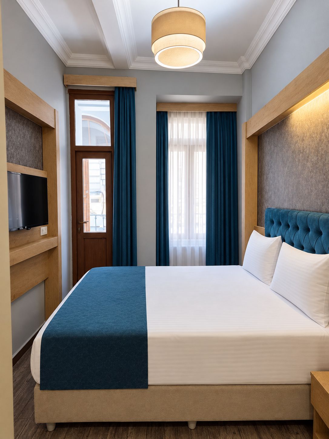 A hotel room with a bed, a television on a stand, a wooden headboard, and a window with curtains.