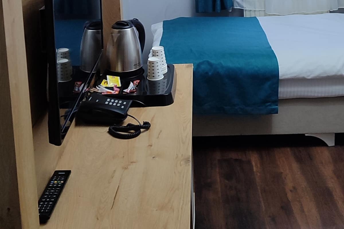 A hotel room with a wooden desk, a flat-screen TV, a bed with blue curtains, and a white ceiling.