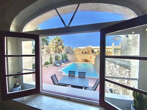 A room with a large arched window overlooking a swimming pool, outdoor seating, and a stone wall.