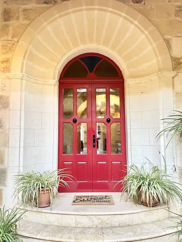 A red double door with glass panels, set within an arched stone entrance, flanked by potted plants.