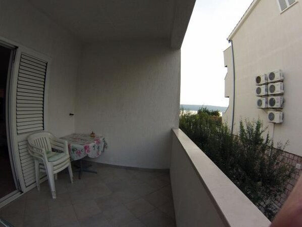 A small balcony with a white chair, a small table with a floral tablecloth, and air conditioning units on the wall.