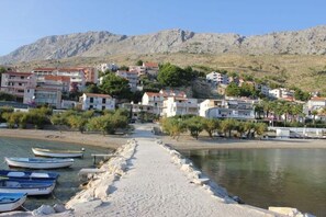 A coastal town with buildings along a sandy beach, boats docked, and mountains in the background.