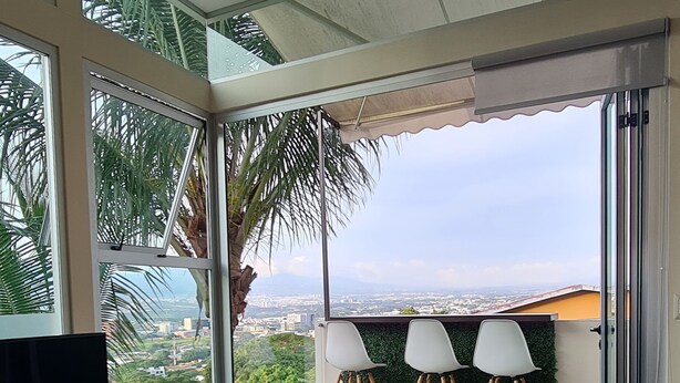 A modern living room with a large TV, a glass coffee table, and a sofa. There are three white chairs and a small table with a plant. The room has large windows offering a view of a cityscape and palm trees. - Cozy Escazú Apartment with great view & location (Escazú)