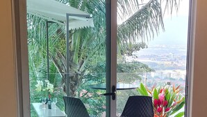 A bedroom with a large window, a bed, a chair, a table, and a view of greenery and buildings. - Cozy Escazú Apartment with great view & location (Escazú)