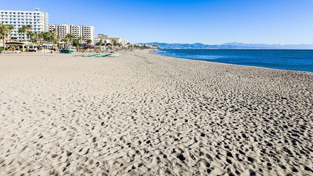 Ein weiter Sandstrand, strahlend blauer Himmel, einige Palmen und Gebäude im Hintergrund.