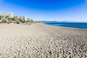 A wide sandy beach with a clear blue sky, a few palm trees, and buildings in the background. - Luxury Apartment at the Marina (Benalmádena)