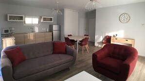 Un salon moderne avec un canapé gris, des fauteuils rouges, une table à manger avec des chaises, un coin cuisine équipé d’un réfrigérateur, d’un four à micro-ondes et de placards, ainsi qu’une horloge murale.
