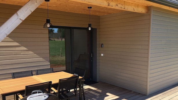 Une terrasse extérieure avec une table et des chaises en bois, un barbecue et une porte-fenêtre coulissante donnant sur un jardin.