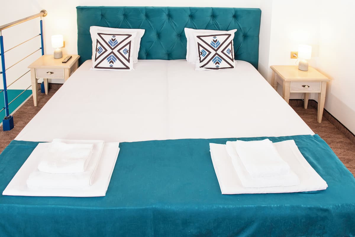 Een tweepersoonsbed met een blauwgroene sprei, twee bijpassende kussens, twee nachtkastjes met lampen, en een blauw hoofdeinde.