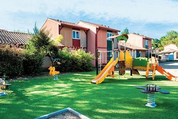 A playground with a slide and climbing structures, surrounded by a well-maintained lawn and a building with a red-tiled roof.