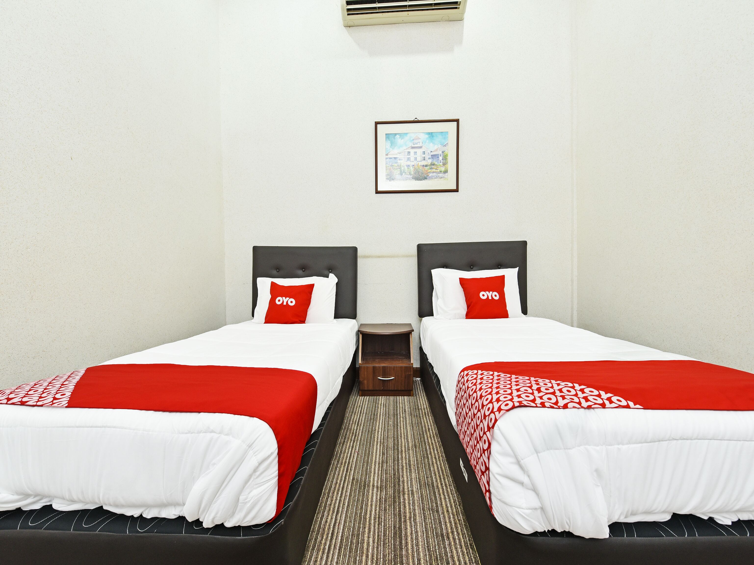 Quarto de hotel com duas camas, cada uma com colcha branca e vermelha, cabeceira escura e mesa de cabeceira pequena.