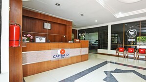 A reception desk with a sign reading "Capital O By OYO," wooden paneling, a clock, a fire extinguisher, and a reception area with chairs and a glass wall.