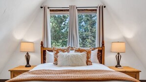Une chambre à coucher avec un grand lit, deux tables de chevet, une fenêtre avec des rideaux et une vue sur les arbres.