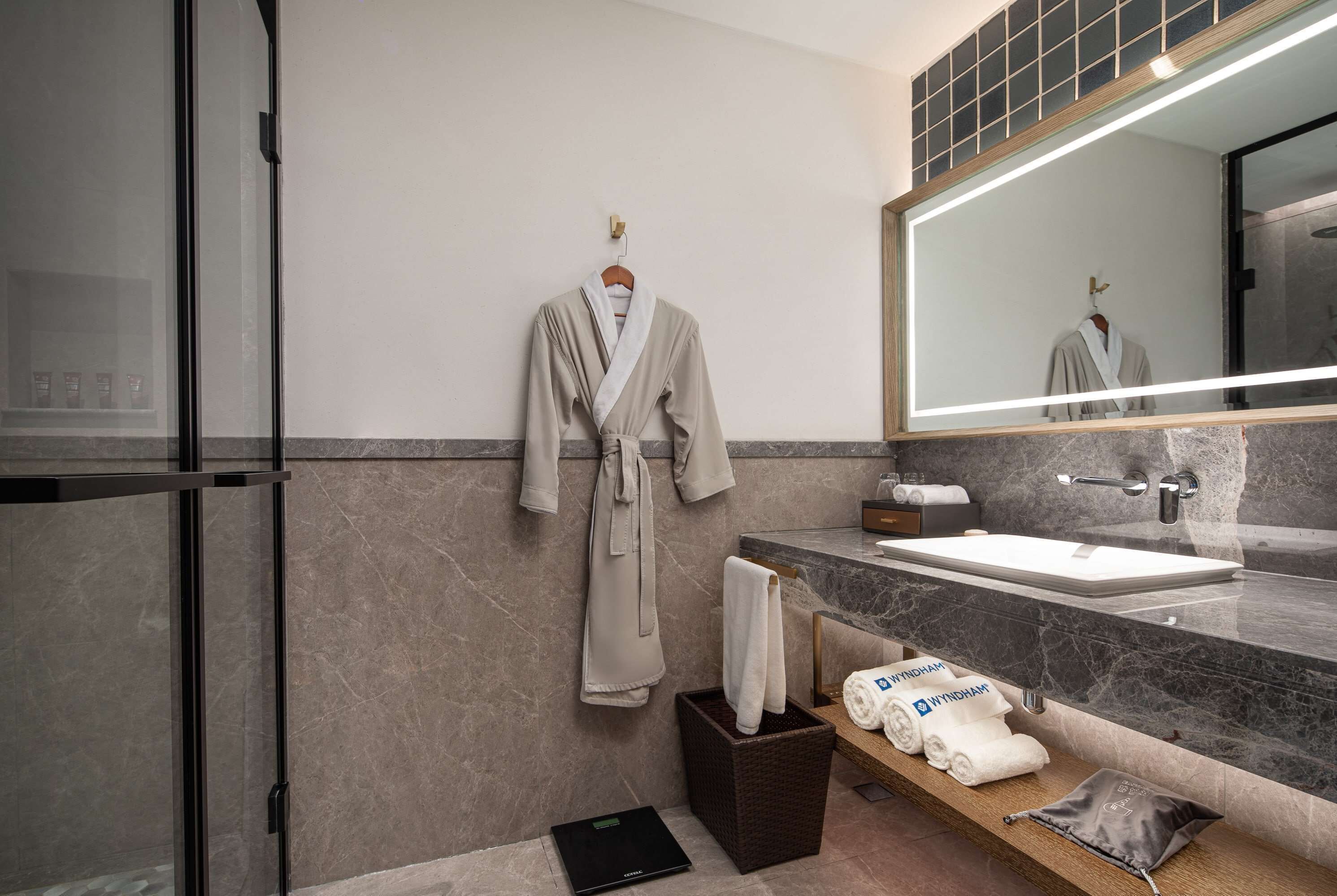 A modern bathroom with a large mirror, a sink, and a robe hanging on the wall. There are towels and toiletries on a shelf.