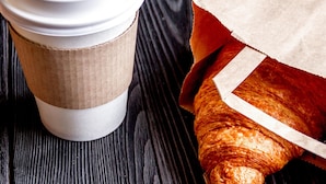 Une tasse de café avec un couvercle et un sac en papier contenant un croissant, le tout posé sur une surface en bois.