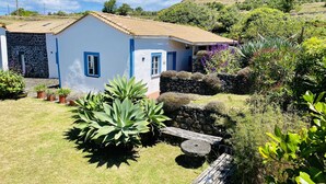 Un bâtiment blanc orné de bordures bleues et surmonté d’un toit en tuiles, entouré d’une pelouse verdoyante et de diverses plantes.