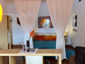 A room with a bed, a table with decorative items, a window with curtains, and a painting on the wall.